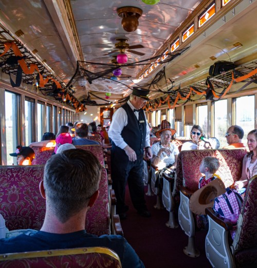 A decorated train interior with passengers, a conductor in a hat, and festive Halloween decorations.