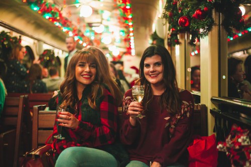 Two women smile while holding drinks on a festive, decorated train filled with holiday decorations and cheerful passengers.