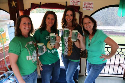 Four women in green shirts hold drinks, smiling and celebrating together in a festive setting.