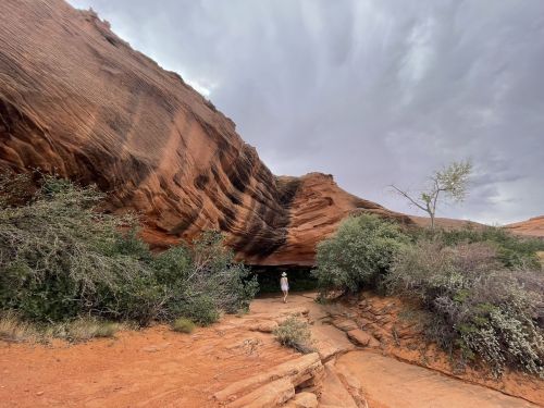 A person stands beneath a large, curved rock formation surrounded by shrubs and a cloudy sky.