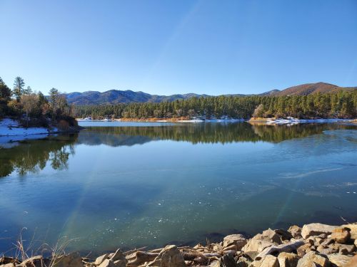 A serene lake surrounded by pine trees and mountains, reflecting the clear blue sky on a winter day.
