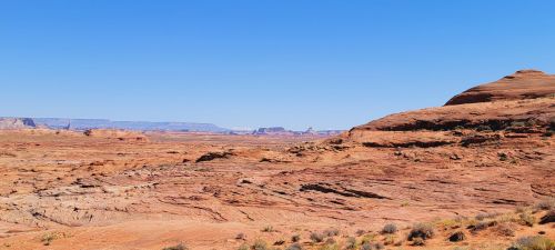 A vast desert landscape with red rock formations under a clear blue sky. Sparse vegetation is visible in the foreground.