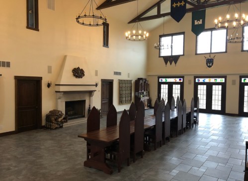 A spacious room with a long wooden table, high ceilings, and large windows, featuring a fireplace and decorative banners.