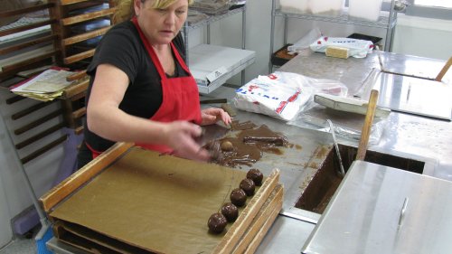 A woman in an apron shapes chocolate truffles on a countertop in a confectionery kitchen.