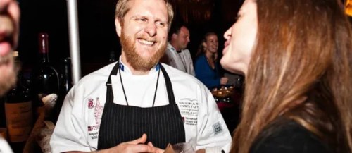A smiling chef in a white coat interacts with a woman at a lively event, with people and wine bottles in the background.
