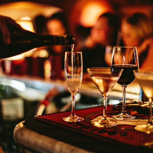 A hand pours champagne into a flute, surrounded by various glasses of cocktails and wine on a bar counter.