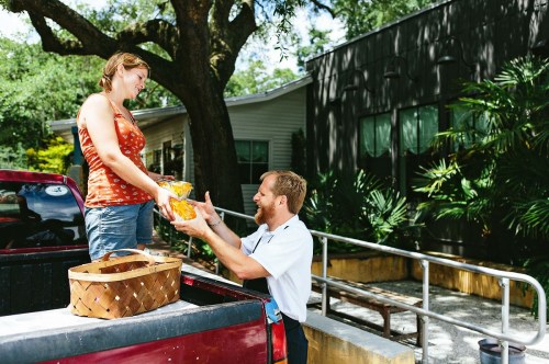 A woman hands a basket of produce to a man sitting in the back of a truck, surrounded by greenery and a building.