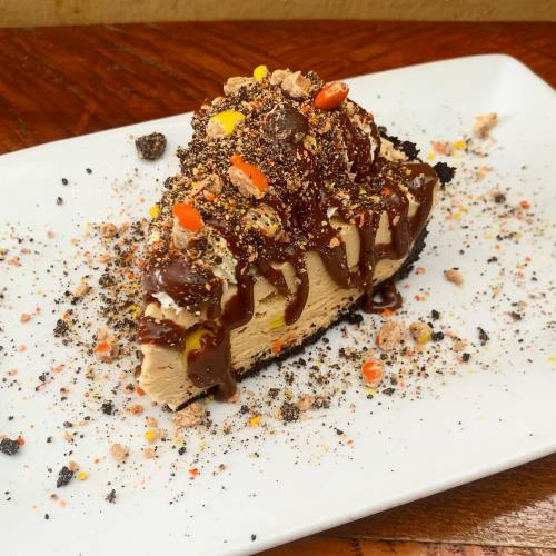 A slice of chocolate peanut butter pie topped with crushed candy, chocolate drizzle, and cookie crumbs on a white plate.