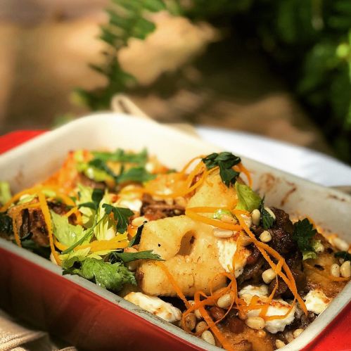 A colorful dish featuring layers of vegetables, herbs, and toppings in a red baking dish, set against a natural background.