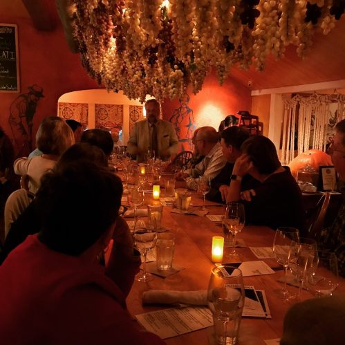 A dimly lit dining room with a long table, guests seated, and a speaker in front, surrounded by decorative grapevines.