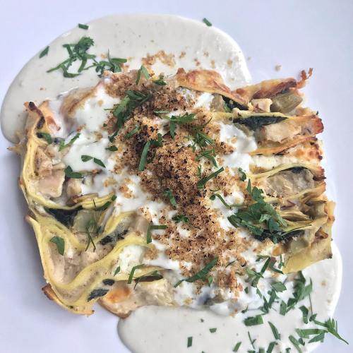 A savory dish featuring layered vegetables topped with creamy sauce, herbs, and crispy breadcrumbs on a white plate.