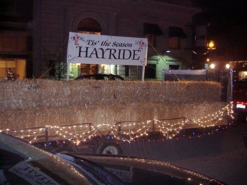 A festive hayride trailer decorated with lights and a sign reading "Tis' the Season" in a nighttime setting.
