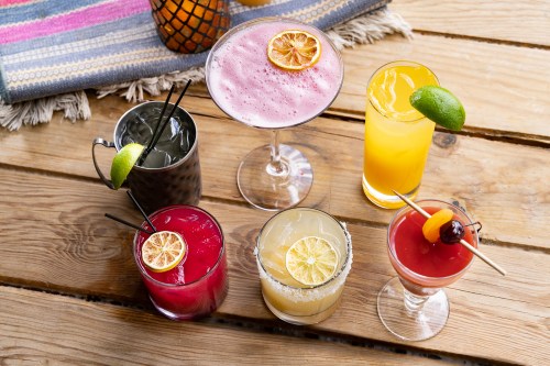 A variety of colorful cocktails on a wooden table, featuring garnishes like lemon and lime slices.