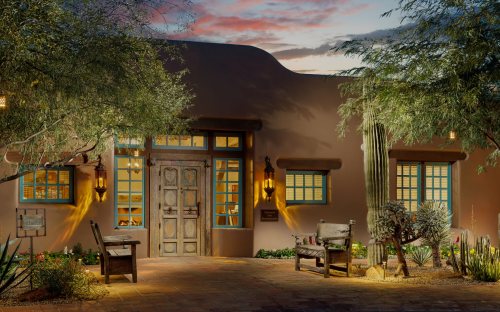 A charming adobe-style building with wooden doors, blue window frames, and desert landscaping at sunset.