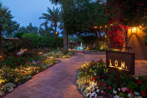 A beautifully lit pathway lined with colorful flowers leads to a pool area, with palm trees and lush greenery in the background.