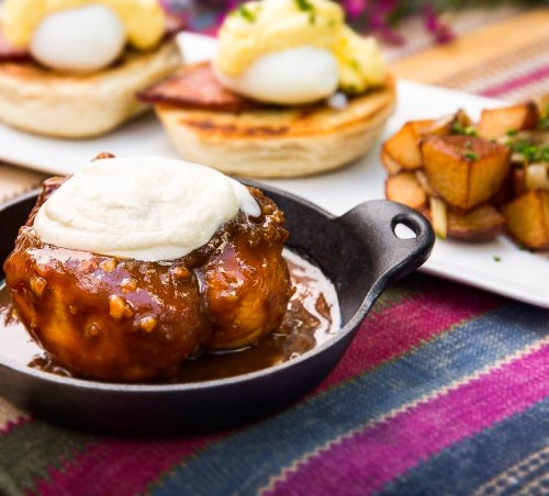 A close-up of a dessert in a skillet, topped with cream, alongside a plate of eggs Benedict and roasted potatoes.
