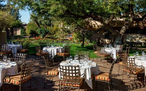 Outdoor dining area with tables set for a meal, surrounded by lush greenery and colorful flowers.