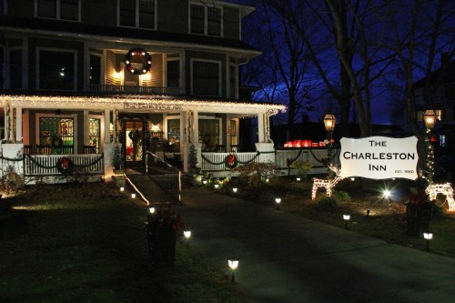 A beautifully lit inn at night, decorated for the holidays, with a sign reading "The Charleston Inn" and festive lights.