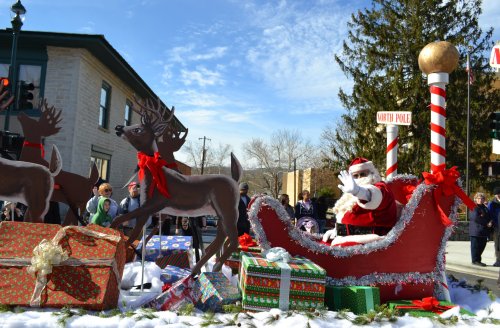 Santa waves from a festive sleigh surrounded by reindeer and colorful presents at a holiday parade.