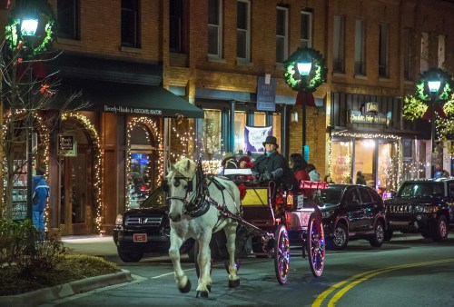 A horse-drawn carriage rides through a festive, illuminated street lined with shops and holiday decorations.