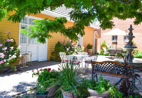 A sunny garden with colorful flowers, a fountain, and outdoor seating surrounded by lush greenery and a yellow building.