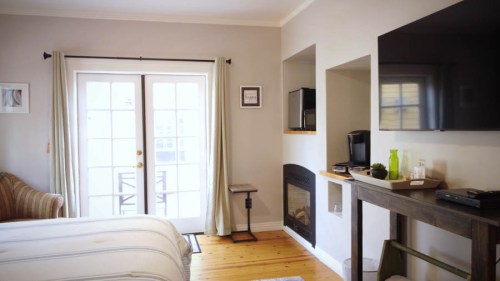 Cozy bedroom with a fireplace, TV, and access to a patio through double doors. Light-colored walls and wooden floors.