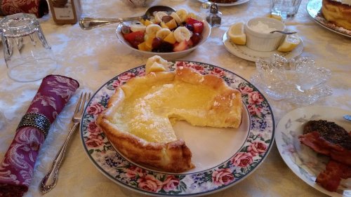 A plate with a slice of baked custard, surrounded by fruit salad, lemon wedges, and other breakfast items on a table.