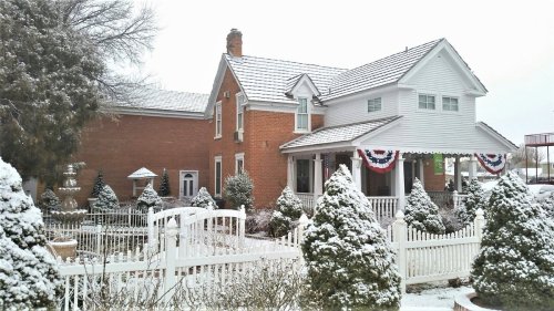 A snow-covered house with a white picket fence, decorated with red, white, and blue bunting, surrounded by evergreen trees.