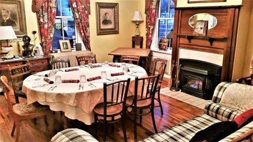 A cozy dining room with a round table set for dinner, a fireplace, and vintage decor, including curtains and framed photos.