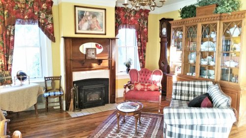 Cozy living room with floral curtains, a fireplace, a plaid sofa, and a wooden cabinet displaying china.