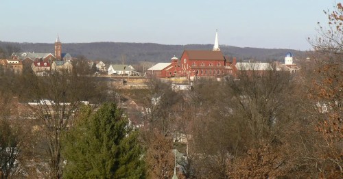 A scenic view of a small town with a prominent church, surrounded by trees and rolling hills in the background.