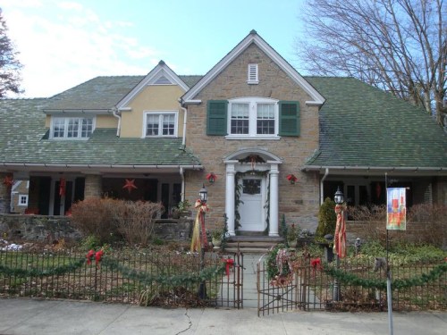 A large, festive house decorated for the holidays, featuring green shutters and a stone facade, surrounded by greenery.