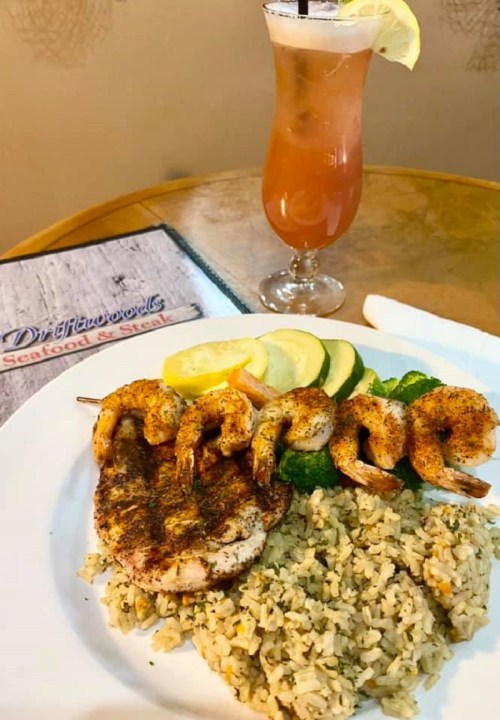 A plate of seasoned shrimp and grilled chicken on rice, with lemon slices, alongside a colorful cocktail.