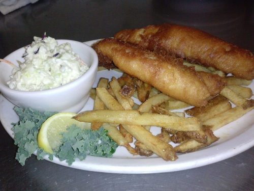 A plate of fried fish fillets, French fries, coleslaw, a lemon wedge, and a garnish of kale.
