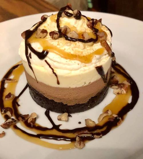 A layered dessert with chocolate mousse, cream topping, drizzled with chocolate and caramel, garnished with nuts.