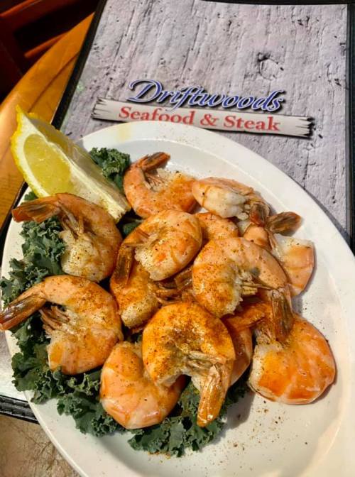 A plate of seasoned shrimp on a bed of kale, with a lemon wedge, next to a menu for Driftwoods Seafood & Steak.