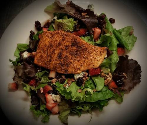 A plate of salad topped with a seasoned salmon fillet, mixed greens, strawberries, pecans, and blue cheese.
