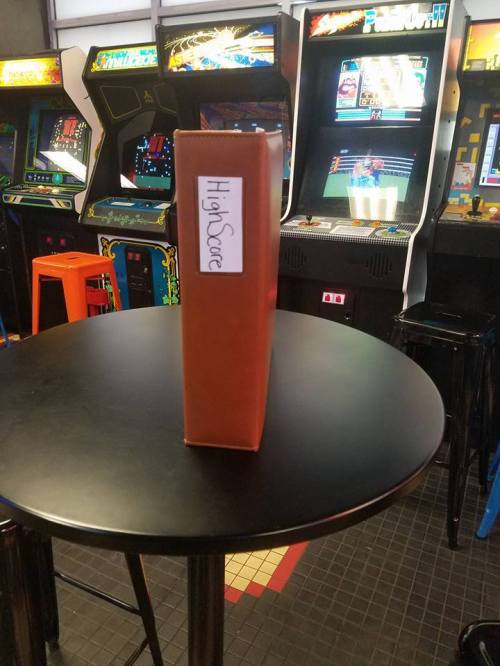 A brown binder labeled "High Score" stands on a black table in an arcade with vintage game machines in the background.