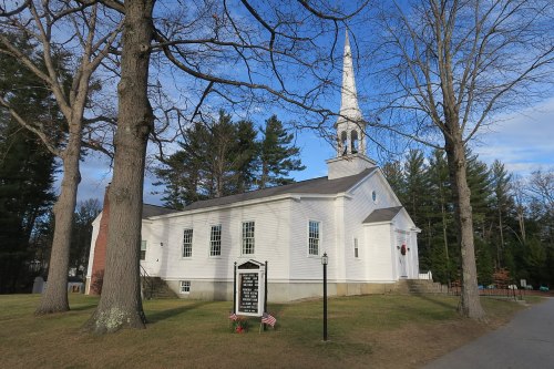 A white church with a steeple, surrounded by trees and a grassy area, featuring a sign in front.