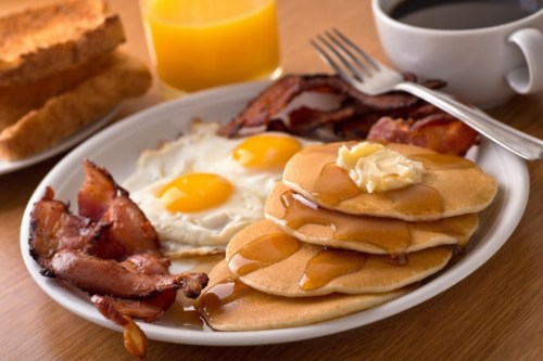 A plate of breakfast featuring pancakes with syrup, bacon, eggs, toast, and a glass of orange juice.