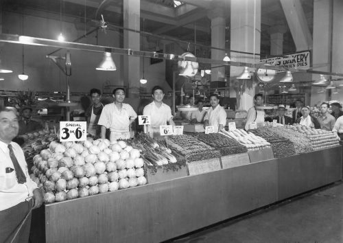 Black and white photo of a bustling market with vendors and fresh produce, including vegetables and baked goods.