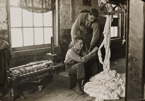 Two young workers in a textile factory, one seated and handling fibers, the other assisting from behind. Natural light from windows.