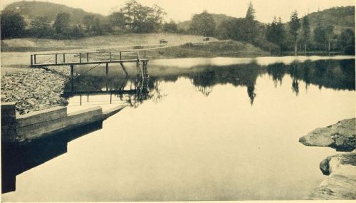 A serene lake scene with a wooden dock, surrounded by trees and hills, reflecting the landscape in calm waters.