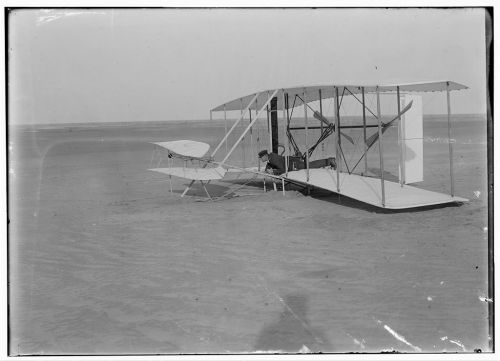 A historic black and white photo of a man preparing to fly a vintage biplane on a sandy landscape.