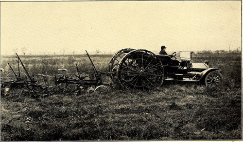 A vintage tractor with large wheels, pulling farming equipment across a grassy field, with a person seated on it.