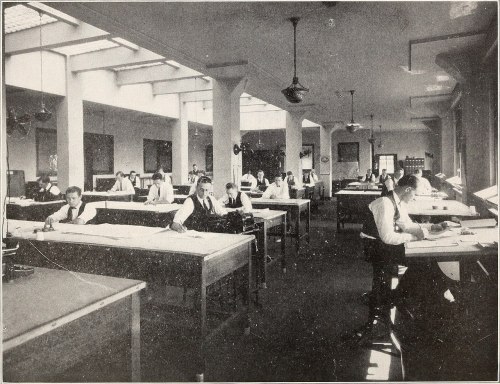 A historic office space with men working at large drafting tables, illuminated by natural light from overhead windows.