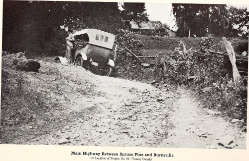 Black and white photo of a rough road with an old car parked beside it, surrounded by trees and a distant building.