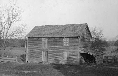 A weathered wooden barn with closed doors, surrounded by a grassy field and bare trees, in black and white.