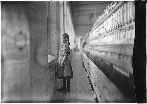 A young girl stands by a window in a textile mill, gazing out, with machinery visible in the background.