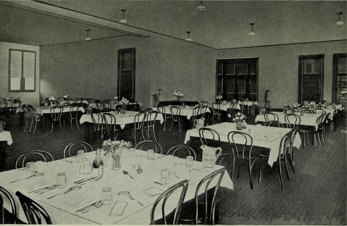 A spacious dining room with tables set for meals, featuring white tablecloths and flower centerpieces.